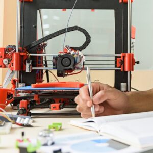 This 3d printer is wonderful. Joyful male engineer is doing research in technology lab. He is sitting and making notes. Man is looking at camera and smiling